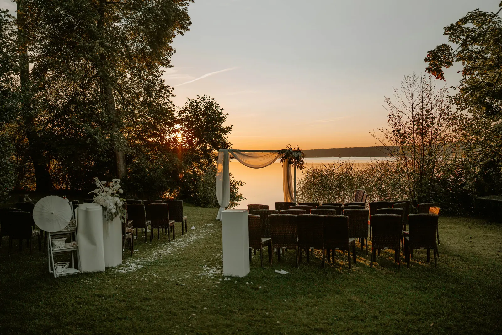 A wedding ceremony set up by a lake at sunset, with chairs facing a draped arch