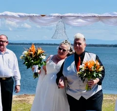 Two brides walking arm in arm with orange bouquets, a bay behind them