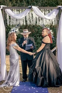 A celebrant in a wide-brimmed hat reading from his notes as two brides hold hands under a draped arch