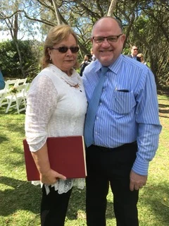 Kelvin at a garden wedding, beside a woman holding the red ceremony folder