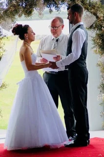 Kelvin reading the vows as a couple hold hands under a floral arch
